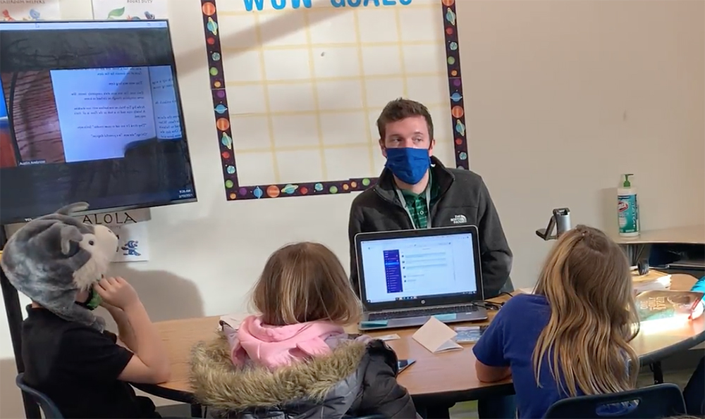 The teacher Austin Ambrose wears a mask while talking to a small group of students sitting at a table.