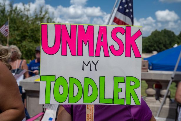 A sign held up at a protest: In neon letters, it says UNMASK MY TODDLER. In the background an American flag is seen. 