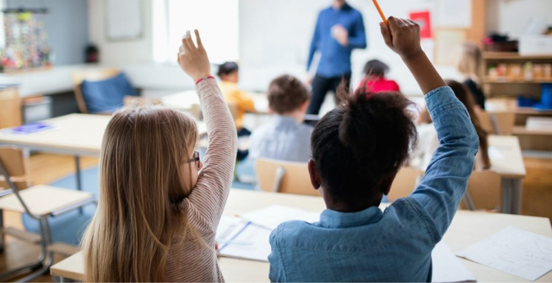 Image of two elementary school students from behind, raising their hands in class