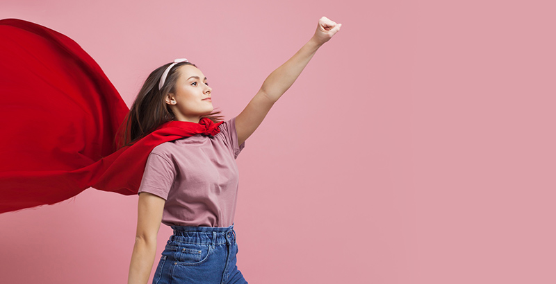 a woman wearing a red cape with fist in the air in front of a pink background