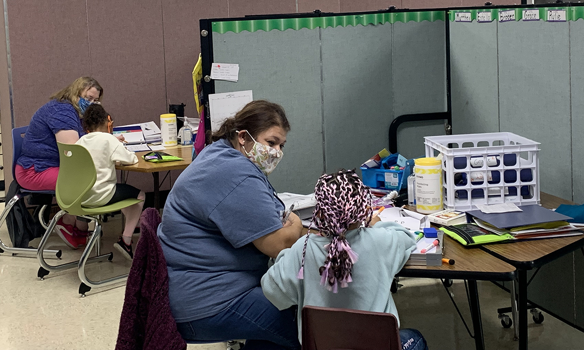 A teacher wearing a mask works with an individual student at a small table in a classroom