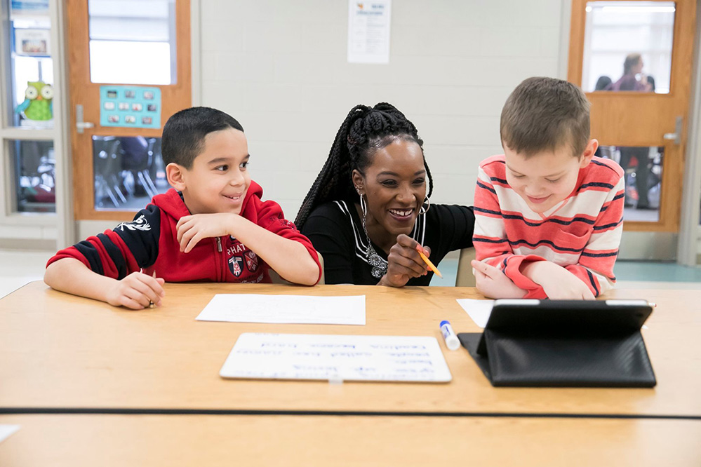 A teacher kneels down to work between two students; all three are smiling