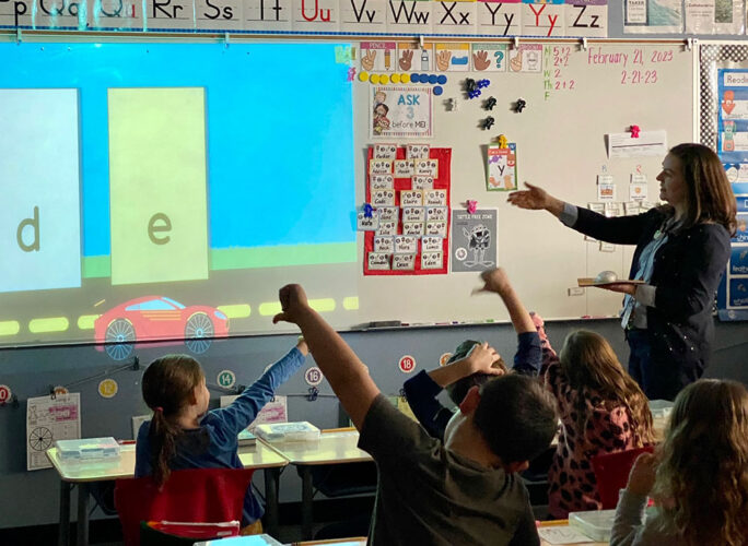 A photo of a teacher at the front of the classroom; many of the students have a hand raised showing a thumbs down