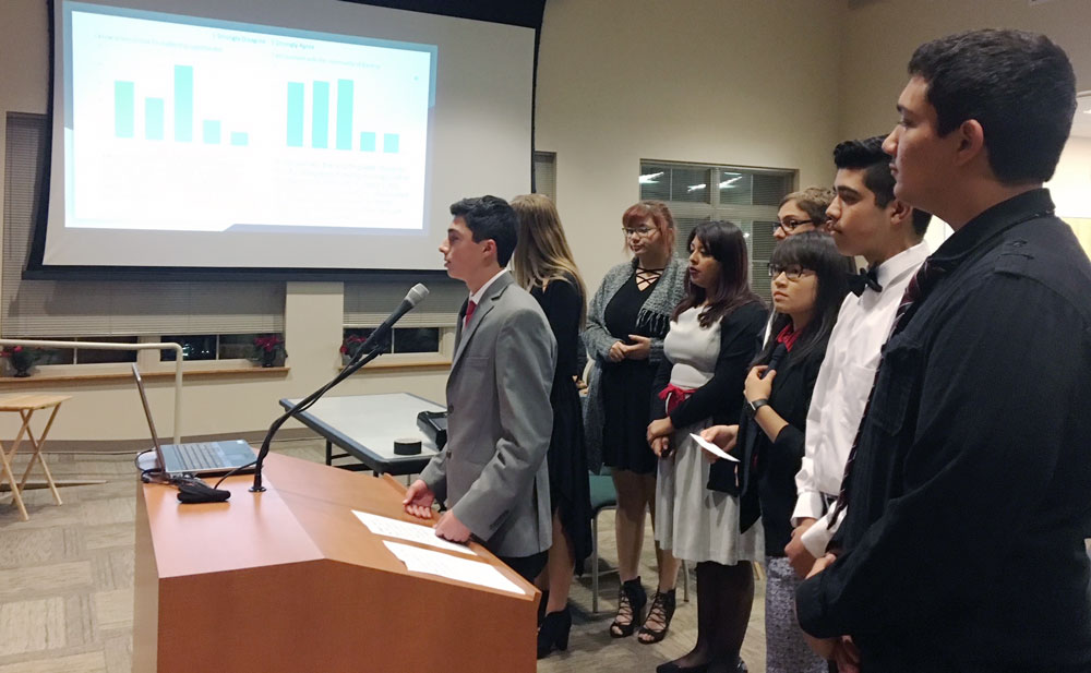 A student speaks at a podium during a city council meeting; several students stand behind looking on