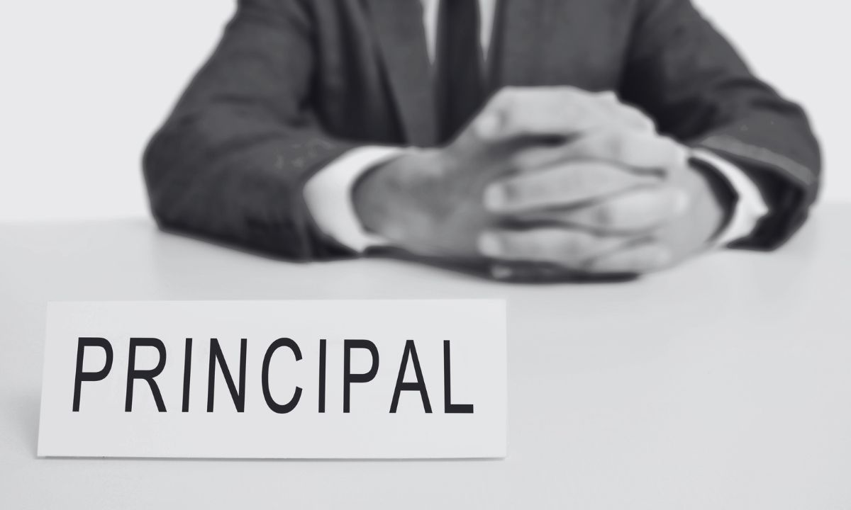 A photo of a man in a suit with a sign in the foreground that says "principal"