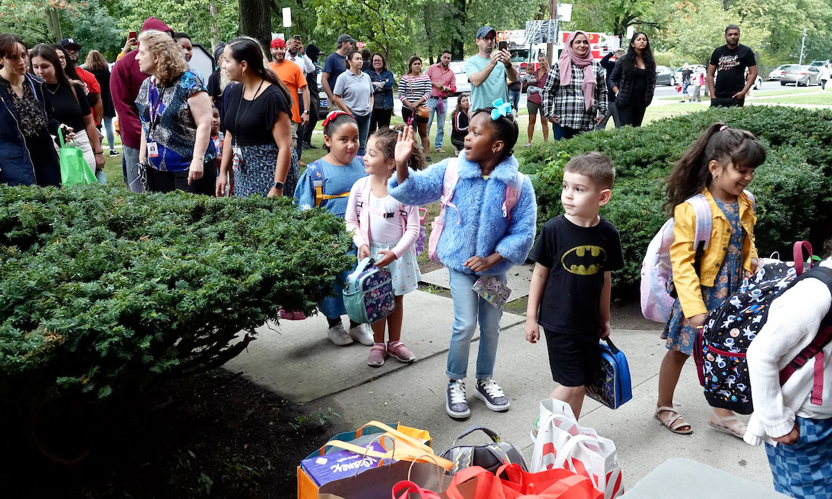 This is a photo of children lined up outside with their backpacks and parents surrounding them.