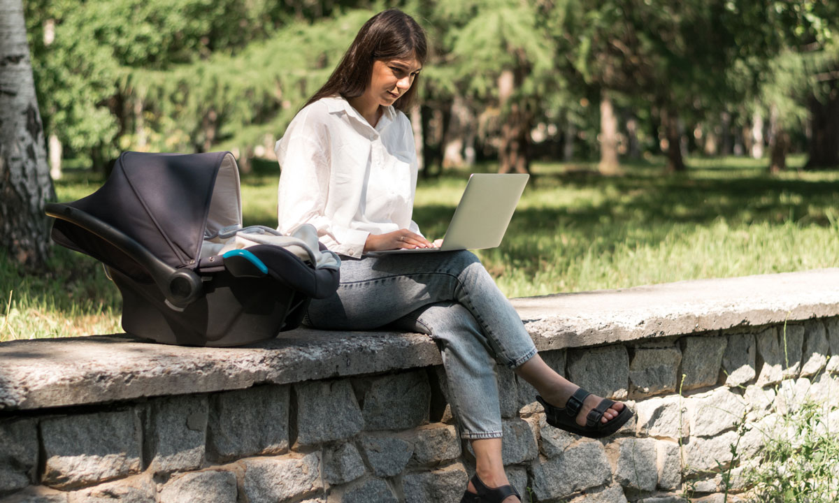 This is a photo of a woman sitting outside on a laptop with a baby in a car seat next to her.