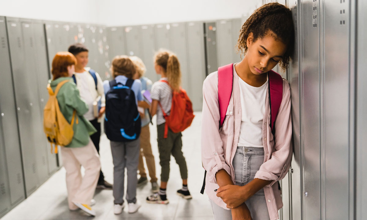 This is a photo of a sad girl standing alone at school lockers with a group of students talking in a circle behind her.