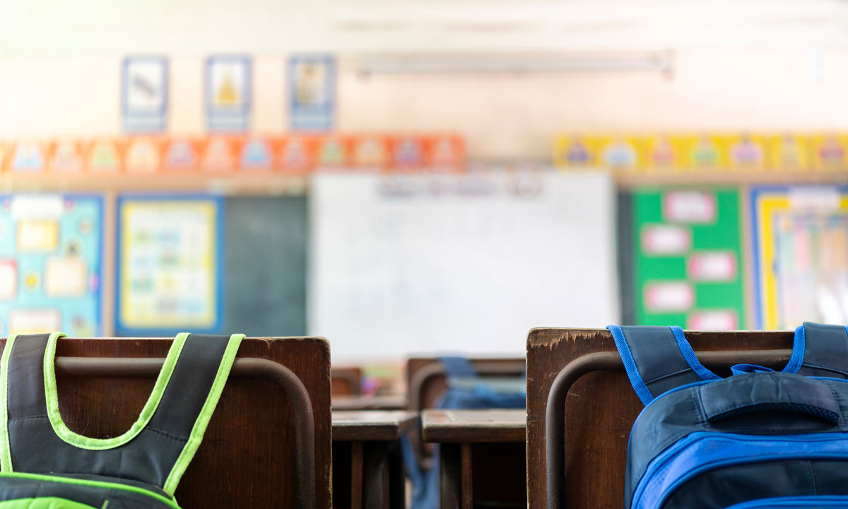 This is a photo of empty desks with backpacks on chairs in a classrooms.